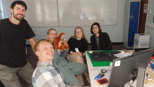 Four students sit round a table in a group. One of them is holding up an orangutan stuffed toy. The orangutan was named Brewster at the summer school and has become a mascot for the CDT in Applied Photonics