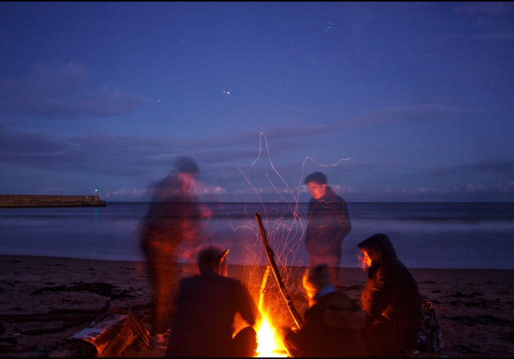 A group of attendees sit around a campfire on the beach in the twilight.