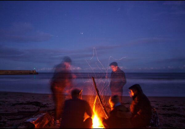 A group of attendees sit around a campfire on the beach in the twilight.