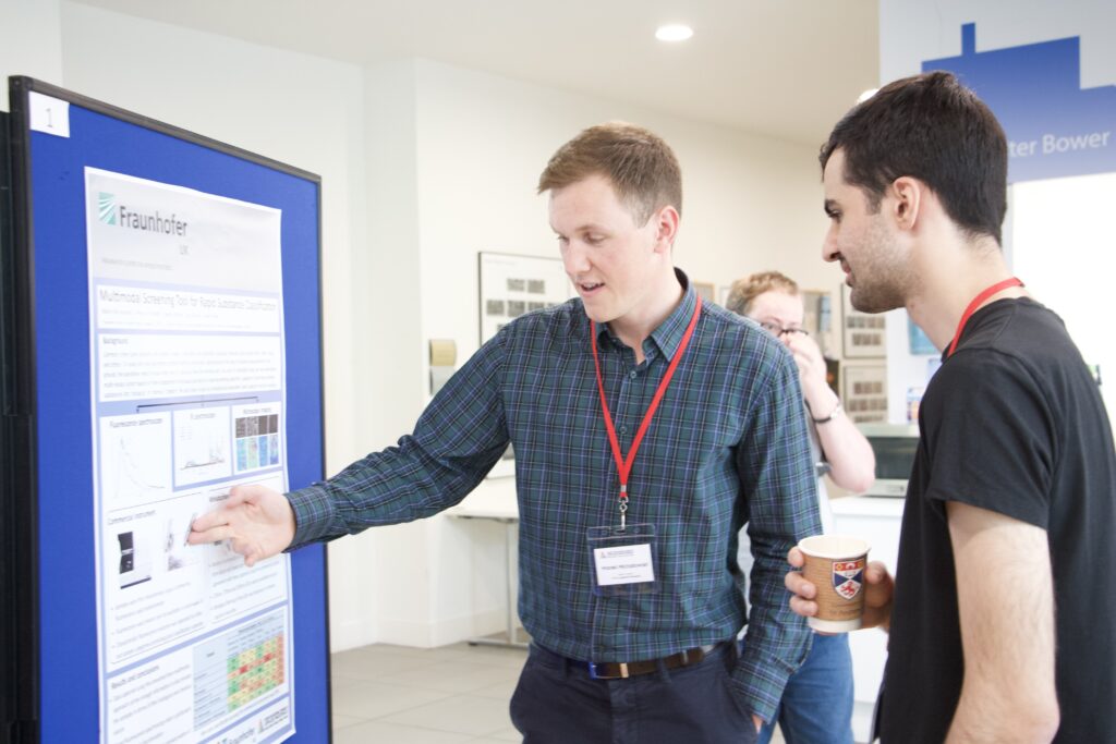 A student is seen presenting his poster to another. The student points to a section of the poster to explain a concept from the poster.