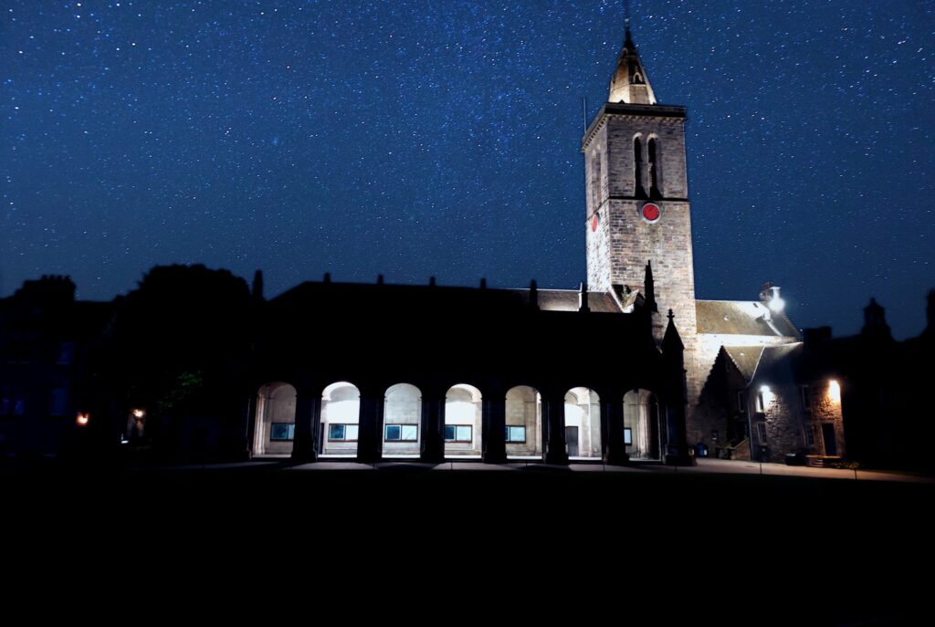 An illuminated building with a tower stands in the foreground. Above an inky blue night sky with stars.